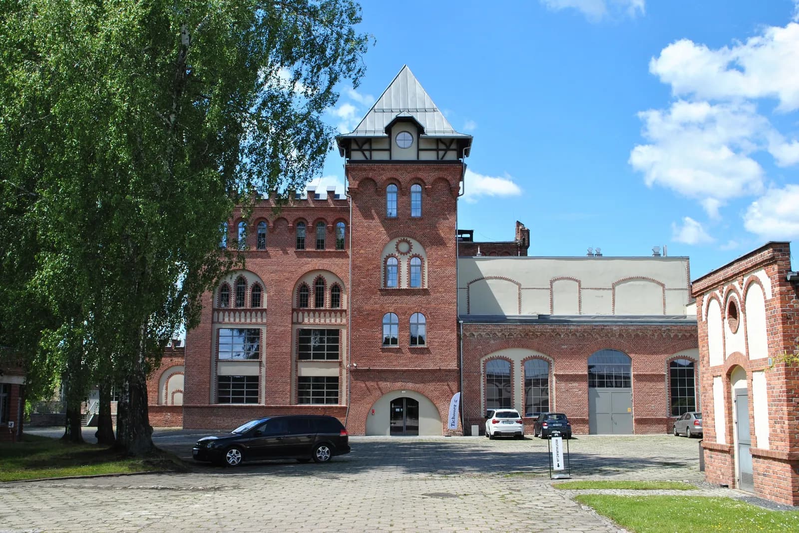 A large red brick building with a tall central tower and many windows, surrounded by green trees and a paved courtyard with parked cars.