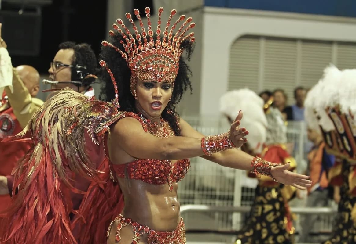 A woman in an elaborate red carnival costume with a feathered headpiece and armbands dances with her arms outstretched, her mouth slightly open.