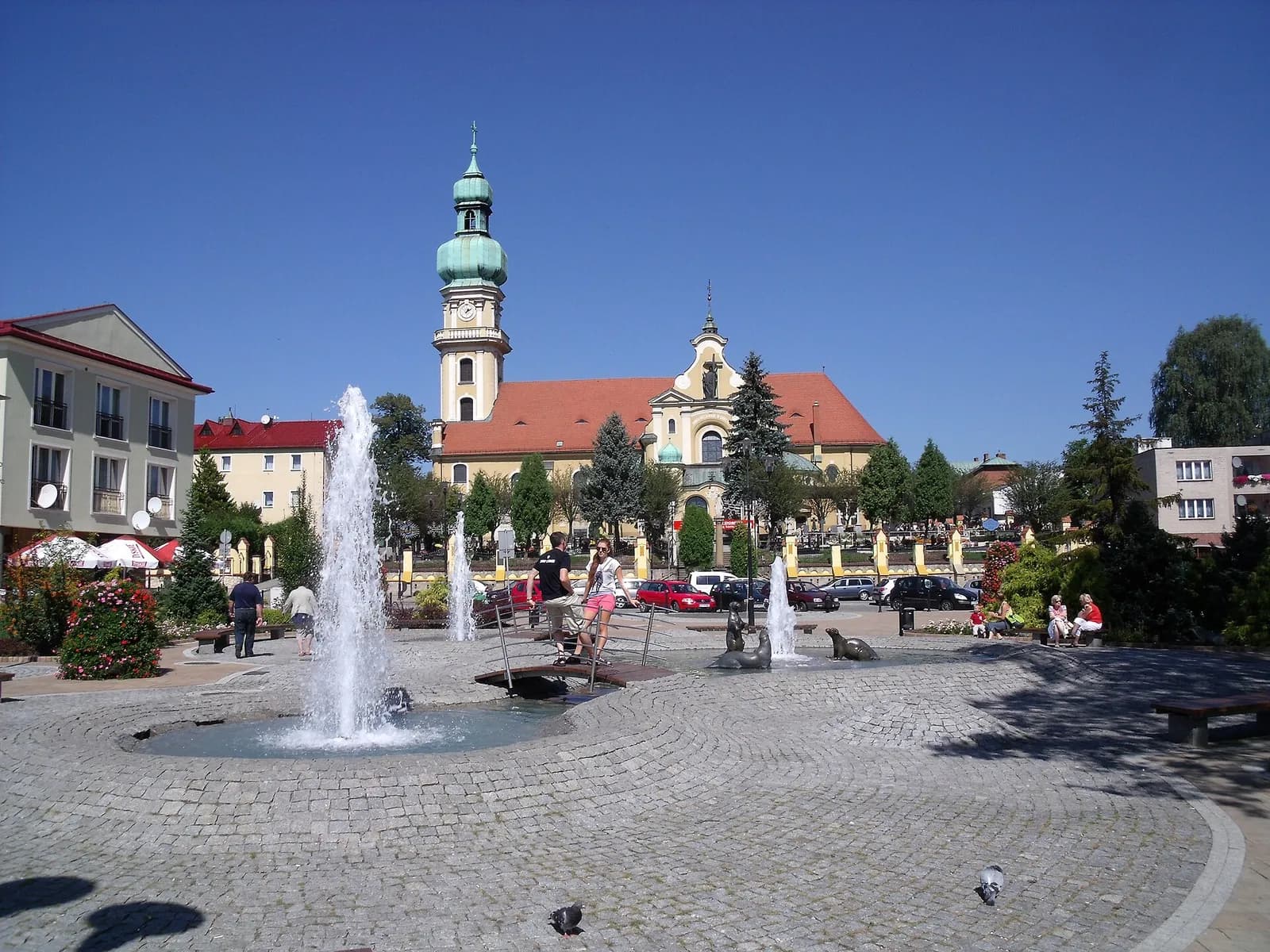 A town square with a large fountain in the foreground and a church with a tall steeple in the background under a clear blue sky.